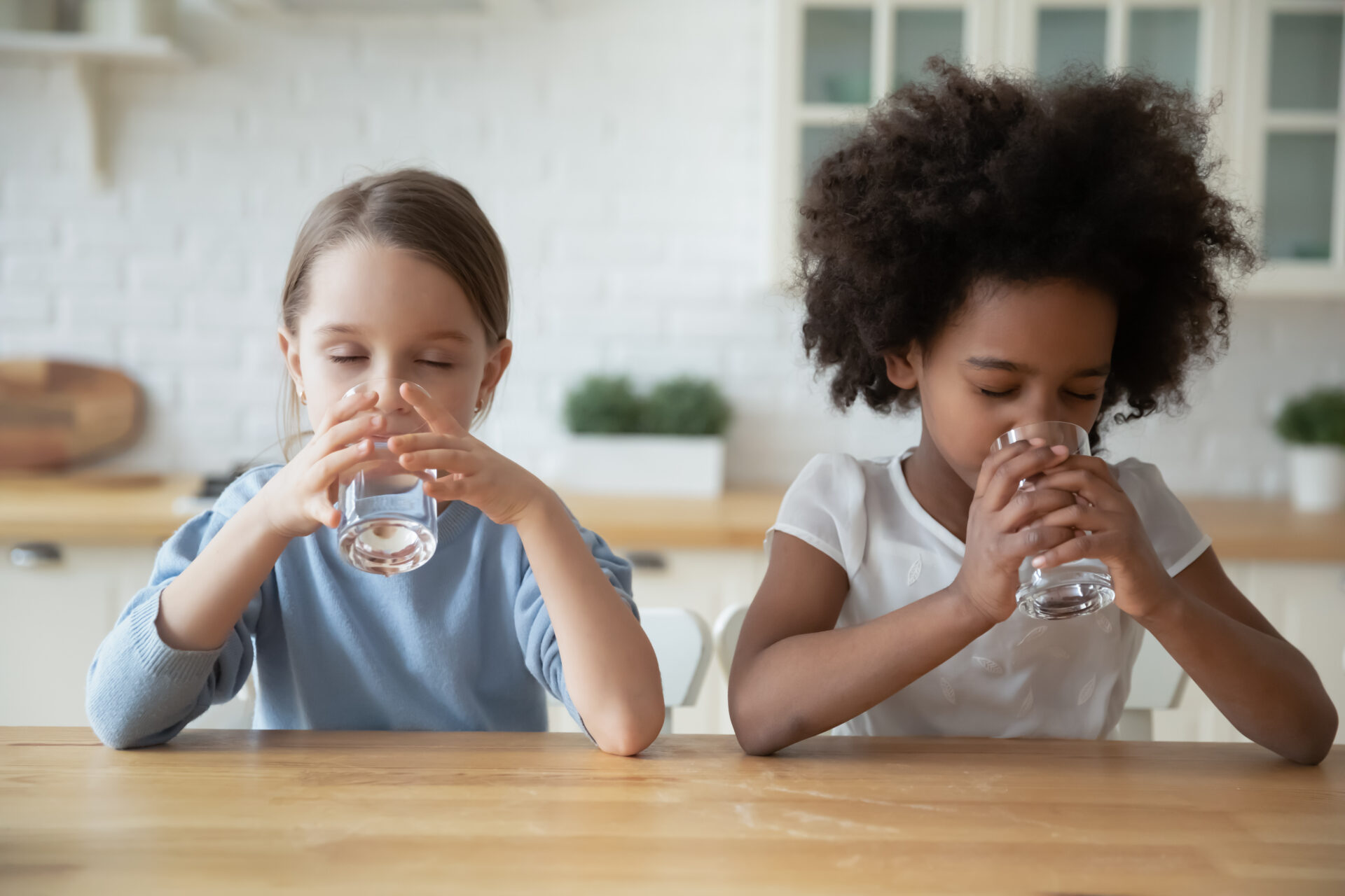 Two multi-ethnic dehydrated little girls reducing thirst drinking natural clean water seated at table in domestic kitchen. Healthy lifestyle, good life habit, daily body organism refreshment concept